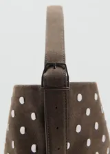 Bolso tipo hobo de cuero gamuzado color marrón, con diseño de tachas metálicas plateadas distribuidas por toda la superficie. Cuenta con una correa de hombro integrada y cierre superior.