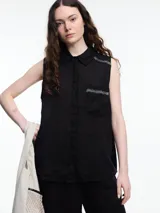 Camisa sin mangas negra, confeccionada en lino, con cuello camisero, cierre frontal de botones, bolsillo delantero y bordado tipo pespunte en color contraste en el frente y canesú de la espalda. Cuenta con cartera abotonada en la parte baja de la espalda y tiene un fit relajado.