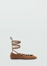 Zapatos chatitos de piel de serraje color marrón, con diseño de tiras cruzadas en el empeine, ojales metálicos dorados y cintas largas para atar alrededor del tobillo. Presentan punta redonda y suela plana.