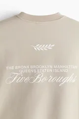 Buzo color beige de corte holgado, con cuello redondo y puños y ruedo acanalados. Estampado pequeño con la frase "New York" y un diseño de hojas en el pecho.