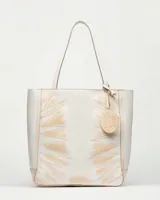 Shopper de cuero color blanco roto con efecto gastado artesanal. Presenta un diseño estampado en tono beige claro con motivos que simulan hojas o rayos, y costuras decorativas en zigzag en el frente. Incluye un dije colgante circular de cuero con logo grabado.