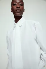Camisa blanca de cuello solapa y manga larga, con detalle de lazada en el mismo tejido en la parte frontal y cierre con botones dorados.