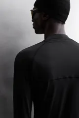 Camiseta negra de running de primera capa, confeccionada en tejido elástico y ajustado al cuerpo. Tiene cuello alto con logo estampado, mangas largas con puños ajustables y costuras planas.