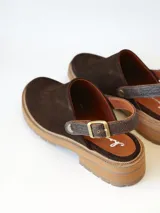 Zueco de cuero gamuzado color chocolate, con diseño de punta cerrada y tira ajustable con hebilla metálica en el talón. Presenta una suela de plataforma dentada en tono marrón claro.