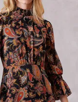 Vestido midi de gasa con estampado de arabescos en tonos cálidos sobre fondo negro. Presenta cuello alto con detalle de punto smock, mangas largas con puños elásticos y volados, y falda con tajo lateral y ruedo con volado. Incluye forro interno y cierre posterior.