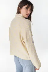 Campera de punto a rayas horizontales en tonos amarillo pastel y lila, con cuello alto, cierre frontal completo y mangas largas con bordado distintivo en la manga.