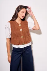 Chaleco tejido color beige con cuello redondo, sin mangas, con cierre frontal de cuatro botones dorados y dos bolsillos frontales.