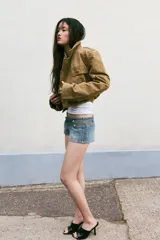 Trench corto de cuello solapa y manga larga, con diseño cruzado y cierre frontal con botones. Presenta bajo acabado en elástico y trabillas en los hombros.