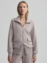 Campera deportiva con cierre frontal, cuello alto tipo chimenea y mangas raglán. Es de corte holgado y presenta bolsillos de parche delanteros. El color es un tono neutro entre gris y rosado (taupe).
