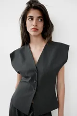 Chaleco de sastre con escote en V, cierre frontal con botones y diseño de hombros caídos. Presenta un estampado de raya diplomática sobre fondo gris oscuro.