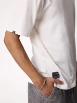 Remera blanca de manga corta y cuello redondo con calce holgado, con una pequeña etiqueta oscura en el borde inferior derecho.