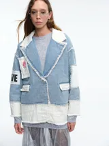 Chaqueta de denim con diseño patchwork, que combina paneles de jean azul claro con secciones de tela blanca acolchada. Presenta solapas amplias, bordes deshilachados, cierre frontal con botones y parches decorativos en las mangas.