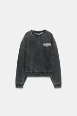 Sudadera de cuello redondo y manga larga, color gris oscuro con efecto lavado. Presenta aplicación de tachas metálicas alrededor del cuello y estampado frontal con el texto "CALIFORNIA USA" en blanco.