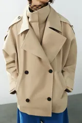 Trench corto oversize color beige con cuello subido de efecto doble, cierre cruzado con botones oscuros y bolsillos de vivo delanteros.