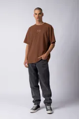 Remera de corte oversize color marrón, con cuello redondo y mangas cortas con hombros caídos. Presenta un bordado minimalista de un joystick en el centro del pecho y una etiqueta aplicada en la parte superior de la espalda.