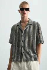 Camisa de calce relajado confeccionada en tejido jacquard con estructura abierta y diseño geométrico. Presenta cuello bowling, manga corta, aberturas laterales en el bajo y cierre frontal con botones.