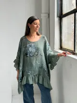 Blusa verde de lino con estampado floral en tonos azules y grises, escote redondo, mangas 3/4 con volados y ruedo asimétrico con volado fruncido.