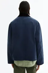 Campera worker corta de sarga de algodón en color azul oscuro, con cuello de pana negro en contraste. Cuenta con cierre frontal oculto por solapa con botones a presión y grandes bolsillos frontales tipo parche.