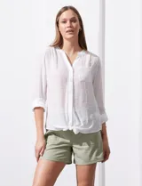 Blusa blanca de gasa marca Zac & Rachel, con cuello a la base, escote en V, abertura central con botones, bolsillo aplicado en el pecho y mangas largas regulables con presilla y botón. Tiene terminación opcional de nudo en el ruedo.
