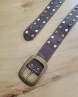 Cinto de cuero marrón con hebilla metálica de estilo envejecido y detalle de tachas metálicas en diferentes tonos en el extremo.