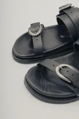 Sandalias planas de cuero negro con diseño de dos tiras ajustables con hebillas metálicas.