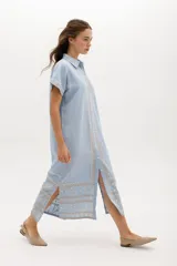 Vestido camisero de lino color lila con bordados rojos en contraste. Presenta un diseño de largo midi con cuello camisero, cierre frontal de botones y mangas cortas. Los detalles bordados recorren la botonadura, los puños y el ruedo de la prenda.