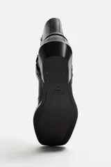 Bota corta negra de cuero con horma cuadrada, tacón bajo y arnés decorativo con remaches plateados a la altura del tobillo.