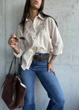 Camisa de color beige con textura rayada vertical, cuello clásico y mangas largas. Presenta un delicado bordado floral en tono sobre tono en la parte superior delantera.