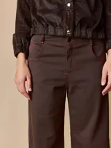 Pantalón de corte relajado color marrón, confeccionado en una mezcla de algodón y elastano. Presenta costuras en contraste color rojo en bolsillos y bragueta, con cierre de botón metálico.