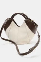 Bolso mini shopper de lona en color crudo con detalles en cuero sintético marrón en los laterales y una manija corta trenzada de mano. Cuenta con costuras contrastantes y cierre magnético.