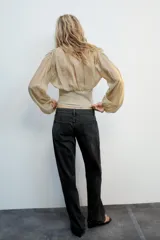 Top beige de manga larga con escote en pico, mangas abullonadas y puños elásticos. Presenta un diseño combinado con tejido fluido en la parte superior y tejido estructurado tipo corsé en la cintura, con cierre frontal de ganchos metálicos.