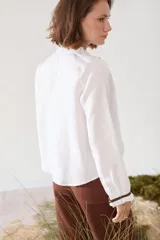 Blusa de lino color marrón con escote en V, cuello con volados y mangas largas con puños fruncidos que presentan detalles en color crudo, al igual que en los hombros. Cuenta con cierre frontal de botones.