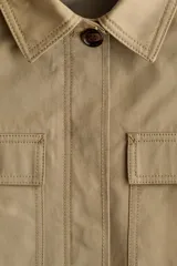 Campera en sarga de algodón color beige, con cuello, cierre frontal con botones ocultos y cordón ajustable en la cintura. Presenta bolsillos de parche con solapa en el pecho y mangas largas.