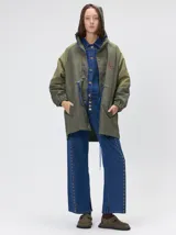 Trench tipo piloto color verde oliva con capucha, cierre frontal con cremallera y cartera de botones. Presenta cordones ajustables en capucha y cintura, puños elásticos, bolsillos laterales amplios y un bolsillo invisible en el pecho con logo bordado.