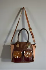 Cartera de cuero combinada en tonos marrones y beige, con detalles en animal print estilo pelo de vaca en los bolsillos frontales. Cuenta con doble asa de mano y herrajes metálicos.