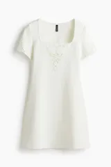 Vestido corto blanco de punto interlock con escote cuadrado, mangas cortas con detalles de encaje y bordado floral en el pecho.