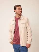 Sobrecamisa de tejido polar color beige, con cierre frontal de botones a presión, cuello clásico y bolsillos frontales con solapa. Presenta un bolsillo adicional con cierre vertical en el pecho y detalles en material técnico.