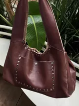 Cartera tipo tote de cuero color marrón con tachas plateadas. Tiene cierre con imán, bolsillo interno con gancho y bolsillo externo con imán.