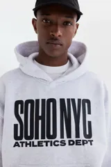 Canguro de felpa gris melange, con capucha de doble capa, bolsillo tipo canguro y estampado frontal con la leyenda 'SOHO NYC ATHLETICS DEPT' en color negro. Presenta hombros caídos y puños y bajo de punto acanalado.