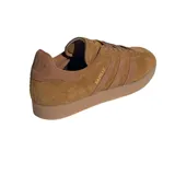 Championes adidas modelo Gazelle, confeccionados en gamuza color marrón con las icónicas tres tiras laterales en un tono más oscuro. Presentan suela de goma color caramelo y el nombre del modelo grabado en dorado en el lateral.
