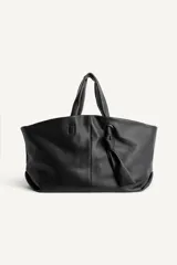 Bolso tipo shopper de cuero negro con textura granulada. Presenta doble asa de mano, cierre magnético y un detalle decorativo de nudo con argolla metálica en el frente.