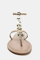 Sandalia plana color beige con tira para el dedo y tira delantera con detalle de abalorios metálicos. Cierre mediante tira con hebilla en el tobillo y acabado en punta redonda.