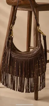 Cartera bandolera de cuero marrón chocolate con flecos largos, tachas metálicas doradas y detalles de textura en relieve. Cuenta con cierre superior y correa ajustable con herrajes dorados.