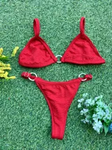 Bikini de dos piezas color rojo, con corpiño triángulo que tiene una argolla central de zamak y bombacha colaless con argollas laterales. La tela es texturada.