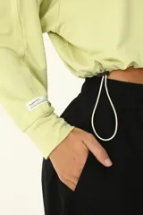 Buzo crop color verde claro, con cuello redondo y mangas largas holgadas. Presenta frunce en el ruedo mediante cordón elástico y tanca.