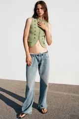 Chaleco de punto en color verde claro, con cuello redondo y diseño sin mangas. Presenta un estilo militar con detalles de alamares, botones decorativos dorados en el frente y bolsillos falsos con tapeta. Cierre frontal con corchetes ocultos.