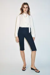 Jeans de tiro medio en denim azul oscuro, con corte capri y pernera entallada. Presenta un diseño con hebilla metálica integrada en la cintura y cierre frontal con cremallera.