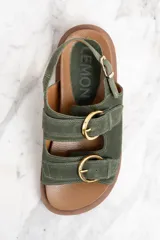 Sandalias de gamuza color verde militar con dos tiras anchas sobre el empeine y una tira trasera, todas ajustables con hebillas doradas. Suela plana color marrón.