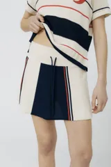 Falda corta de tejido de punto, color crema, con bloques de color azul marino y detalles de franjas verticales en rojo y blanco. Presenta cordón ajustable en la cintura.