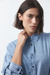 Camisa de jean en tencel con silueta amplia, hombros caídos, cuello clásico y cierre frontal con botones. Presenta ruedo curvo con tajos laterales y pliegue en la espalda.